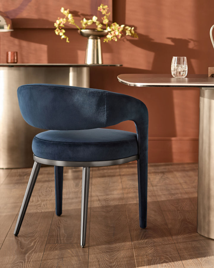 Dark navy velvet dining chair with curved backrest and metal legs, shown in a modern interior setting.