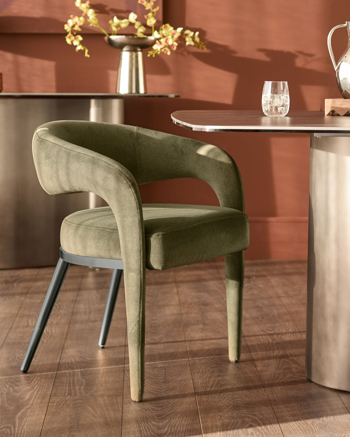 Rhett dining chair in olive green fabric with curved backrest and black metal legs, shown at dining table.