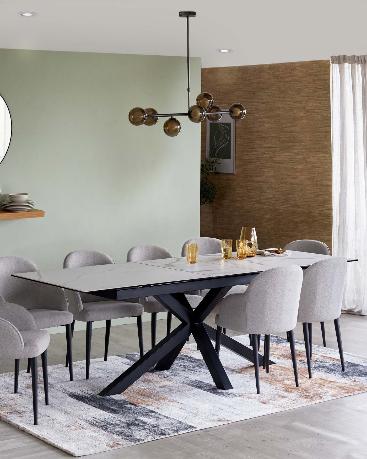 Modern dining room with a black and white marble rectangular table and six grey upholstered chairs with black legs. A patterned area rug under the table provides a contrast with its sandy and grey hues, and a minimalist chandelier with spherical, tinted glass light fixtures hangs above the table.