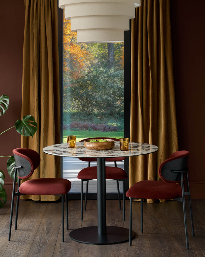A round marble dining table with dark chairs, placed in front of a window with golden curtains and a view to an autumn garden.