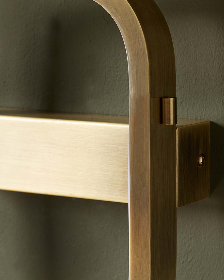 Close-up of a brass towel rail mounted on a textured, dark green wall. The rail is made of bent and rectangular metal pieces.