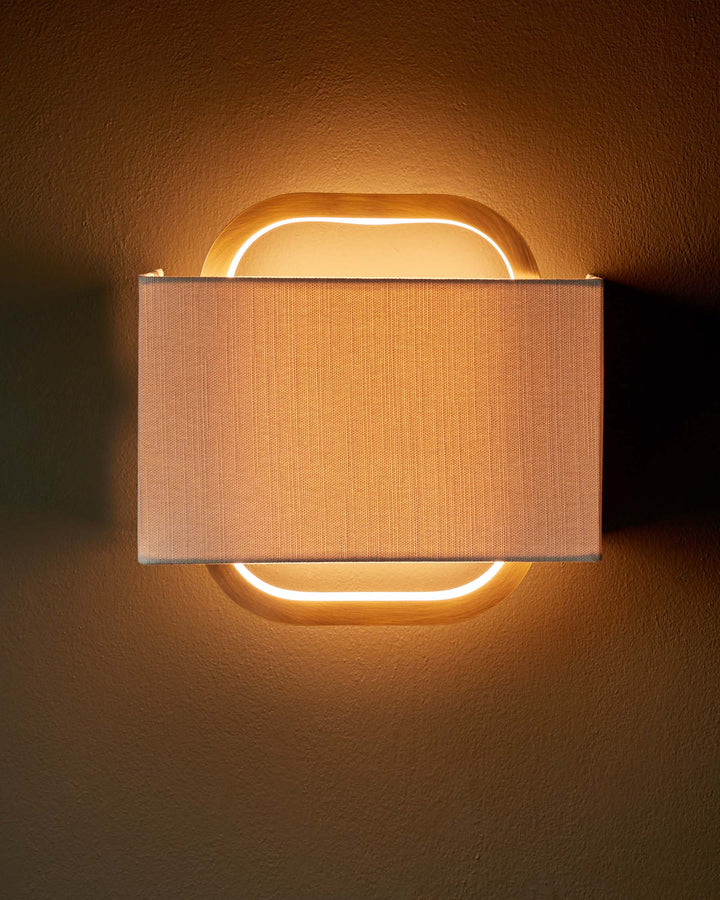 A wall-mounted lamp with a wooden frame and rectangular fabric shade. Warm, inviting light illuminates the textured wall.