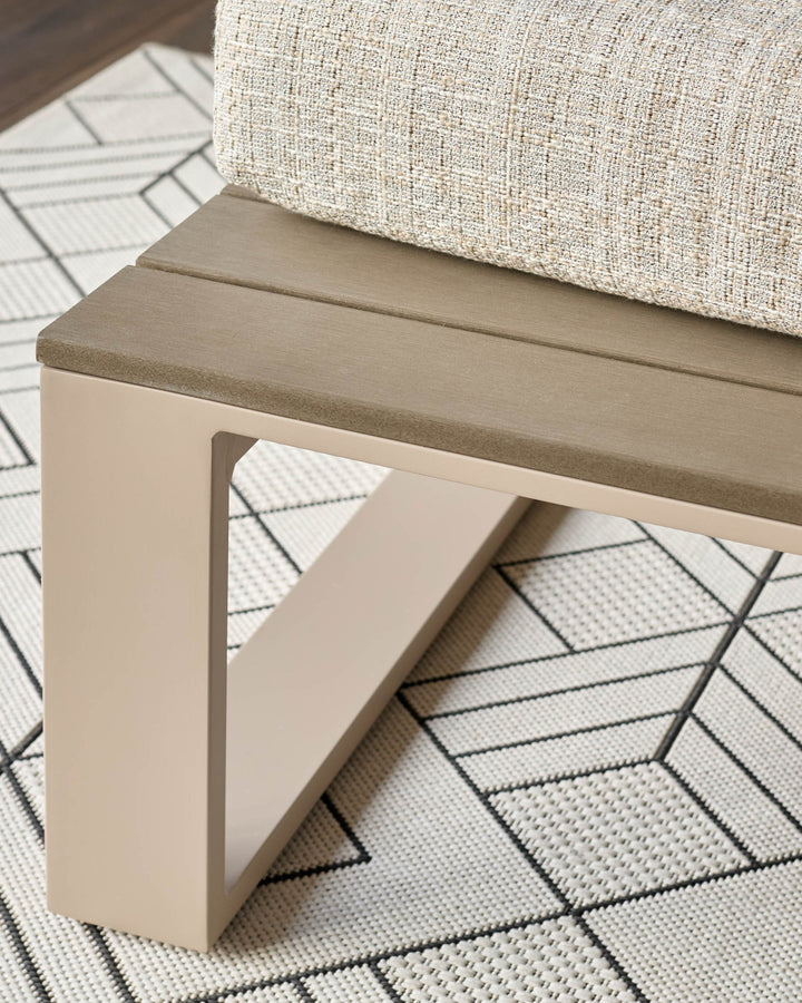 Cream-colored bench with taupe cushioned seat and open geometric base, displayed on patterned rug with cream pillow.