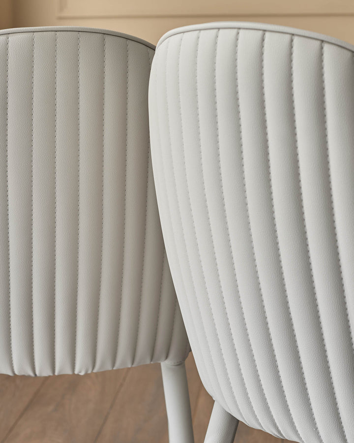 Two light grey faux-leather dining chairs with vertical stitched paneling and curved backrests, shown from a close-up side angle.