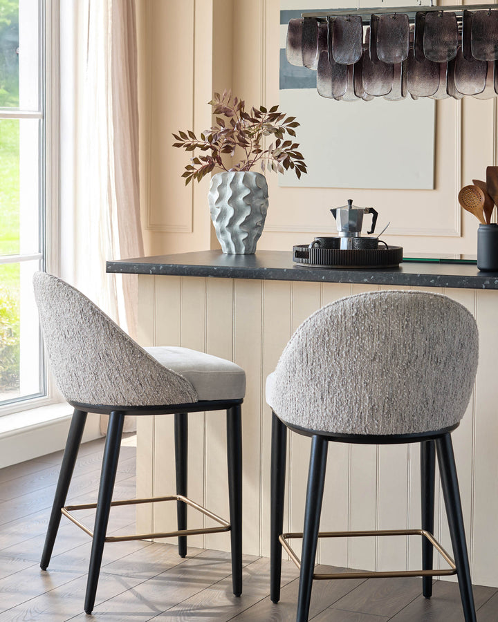 Two stylish bar stools with textured grey fabric, dark legs and brass footrests sit at a kitchen counter.