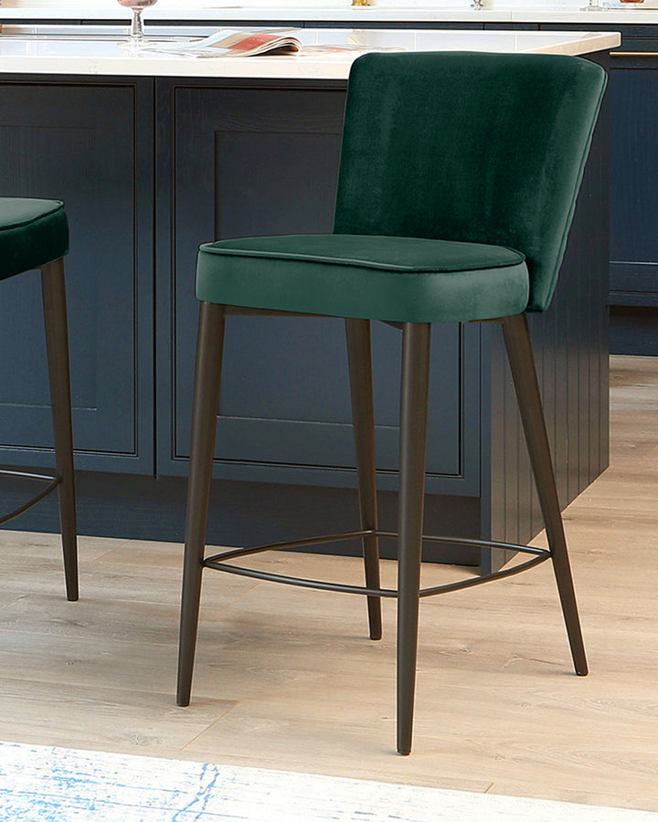 Serena dark green velvet bar stool with wooden legs and metal footrest, shown at an angle in a kitchen setting.