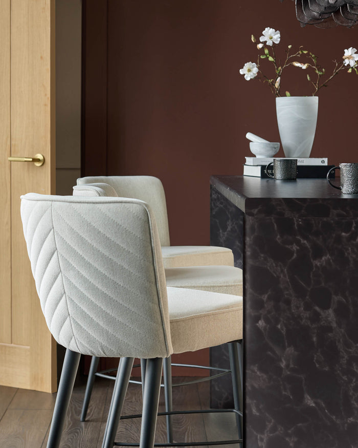 Two stylish, upholstered bar stools with dark metal legs sit beside a dark marble-effect breakfast bar in this interior shot.