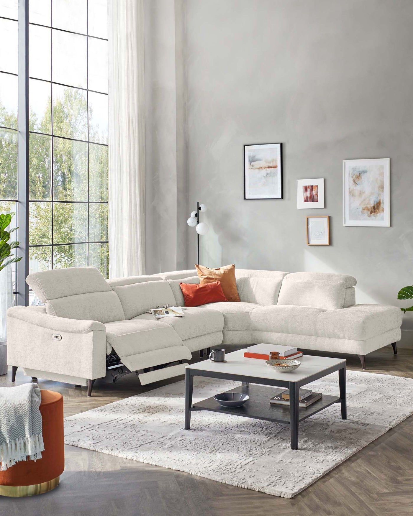 Modern light grey sectional sofa with adjustable headrests, complemented by a terracotta cylindrical side table with a metallic base. In the centre, there is a minimalist black-framed coffee table with a white top, featuring a lower shelf. The set rests on a textured off-white area rug.