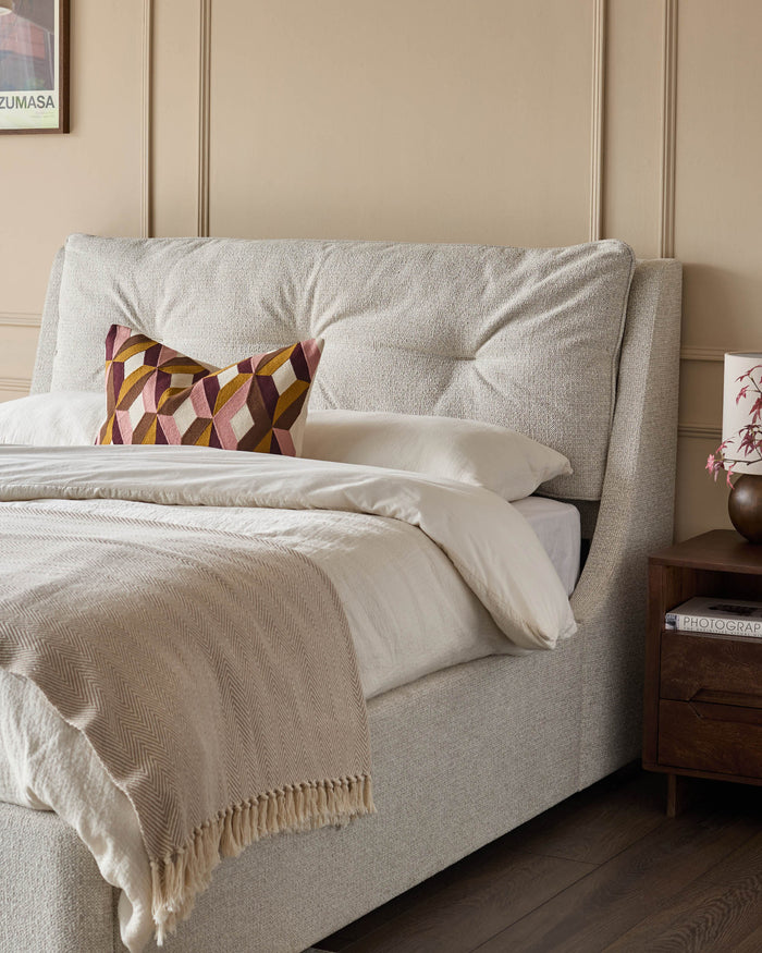 Upholstered bed with patterned cushion and throw, next to a wooden bedside table with lamp and books.