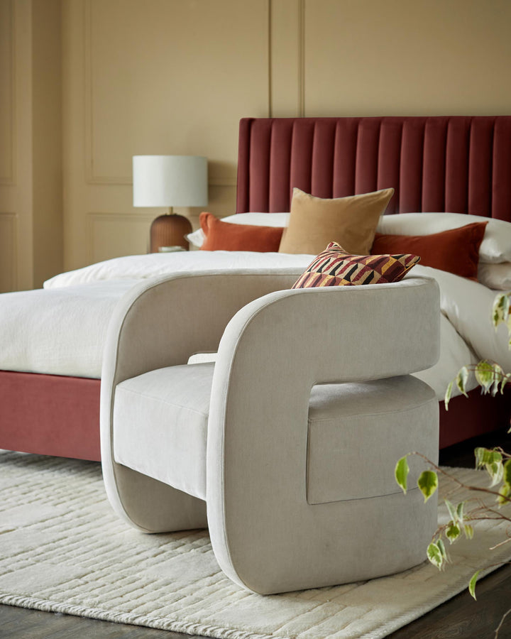 A stylish bedroom setting with a plush armchair in the foreground and a luxe bed with a padded headboard in the background.