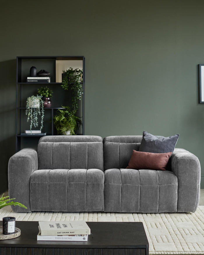 A grey, modular sofa with 2 cushions, on a patterned rug, with a bookcase behind, against a dark green wall.