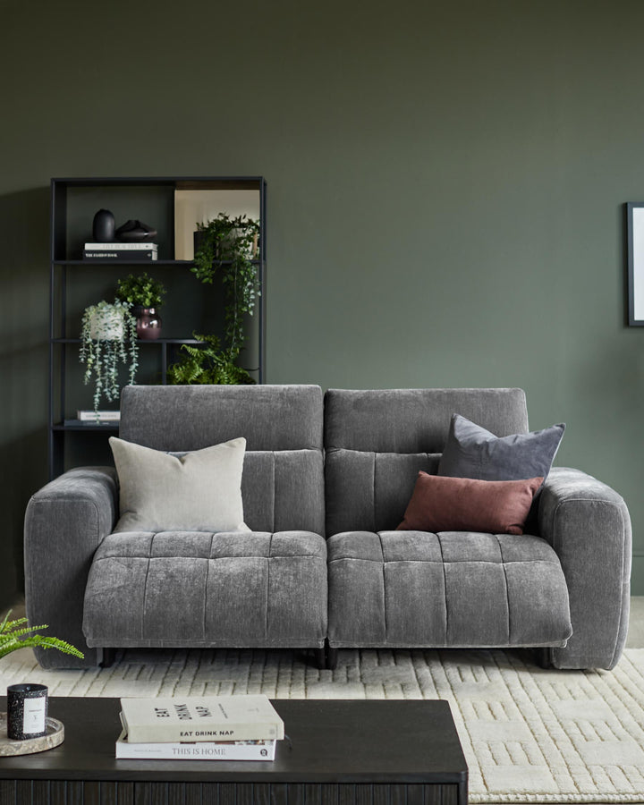 A stylish grey sofa with cushions sits in front of a dark green wall and shelving unit, creating a cosy living space.
