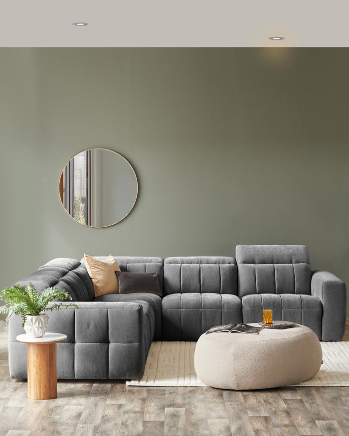 A plush grey sectional sofa with patterned cushions, a round mirror, a light wood side table, and a soft, beige pouf.