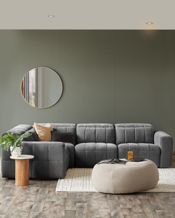Gray sectional sofa with decorative pillows, a round wooden side table, and a soft, upholstered ottoman on a patterned rug.
