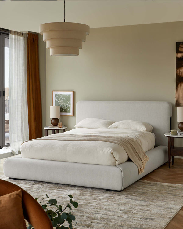 A bedroom scene with a light grey upholstered bed, bedside tables, and layered pendant light.