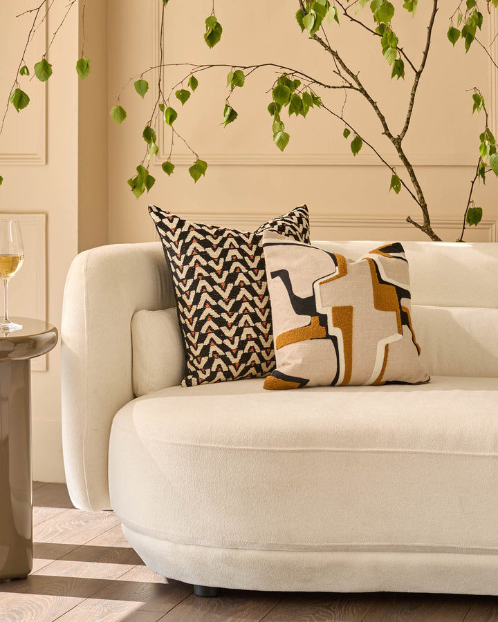 Camille cushion in natural displayed on a cream curved armchair with decorative foliage and a side table.