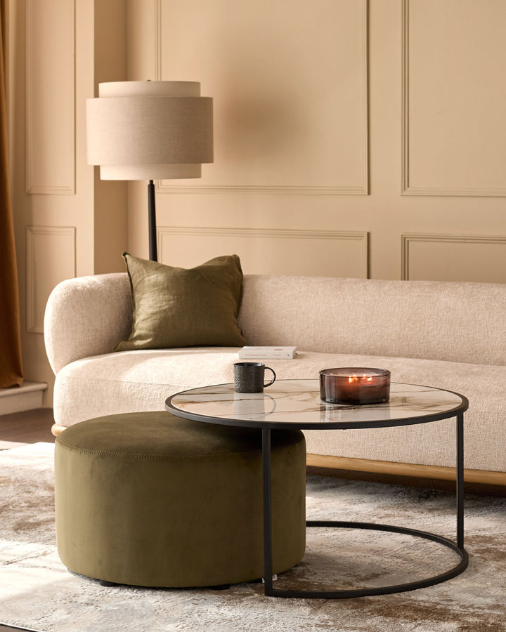 Thea black round coffee table with olive green pouffe in a modern living room setting with cream sofa and lamp.