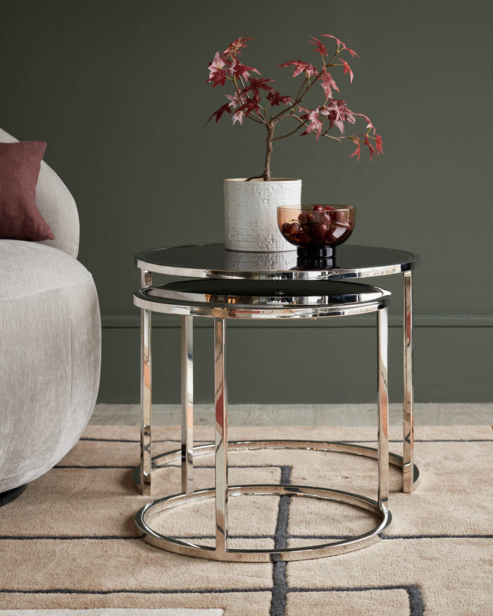 A set of two chrome nesting tables with black glass tops. On top are a vase with branches and a bowl of fruit.
