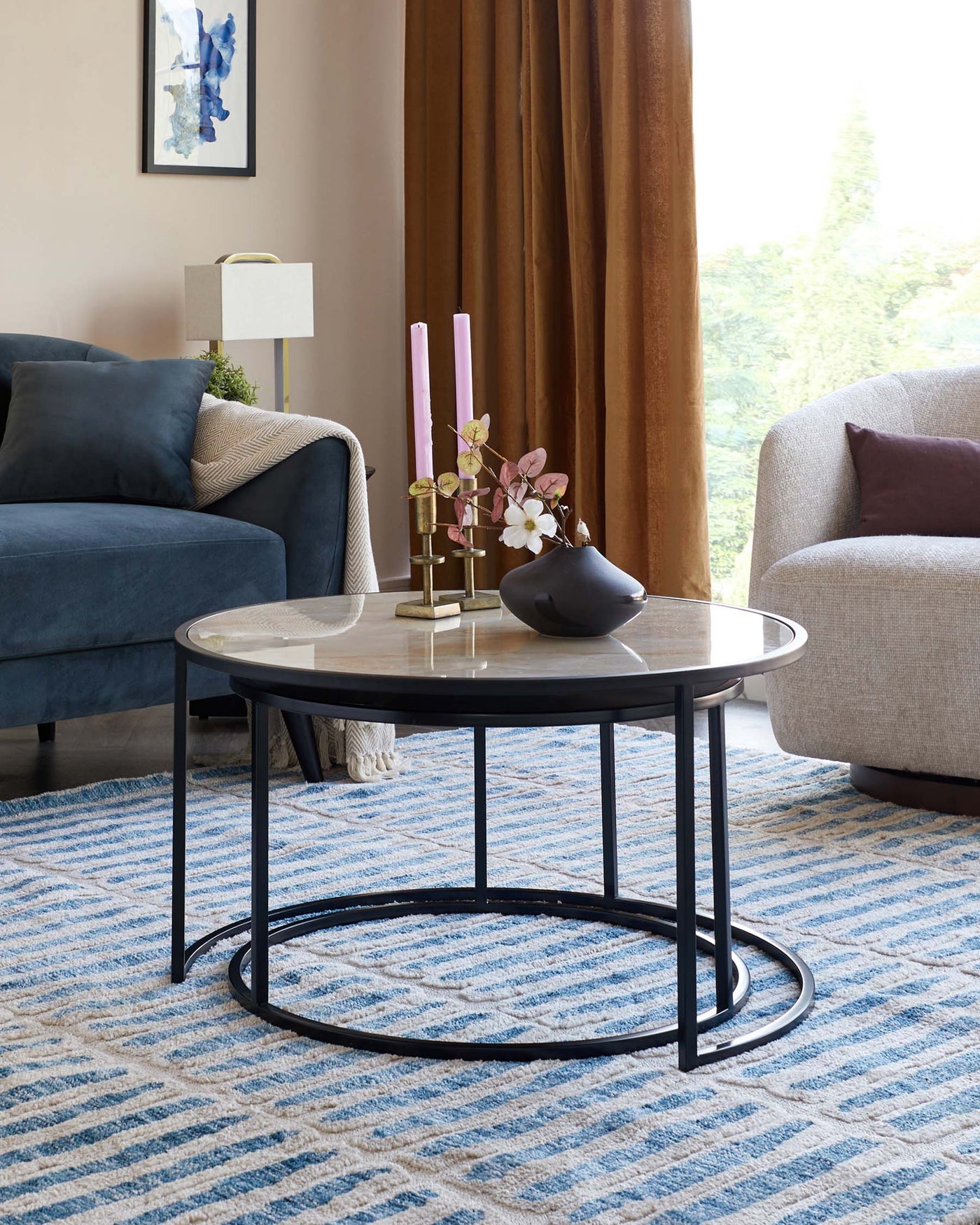 Round marble-top coffee table with a black metal frame, positioned on a blue and white patterned area rug, flanked by a dark-blue upholstered sofa and a light-beige armchair.