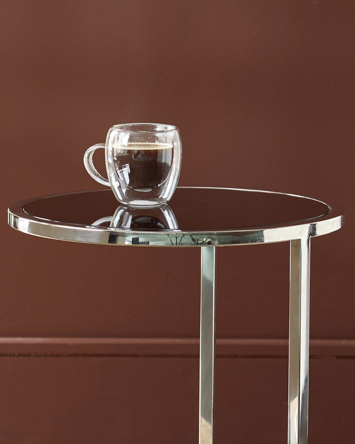 Thea smoked mirror tray table with stainless steel frame holding a double-wall glass mug against brown background.