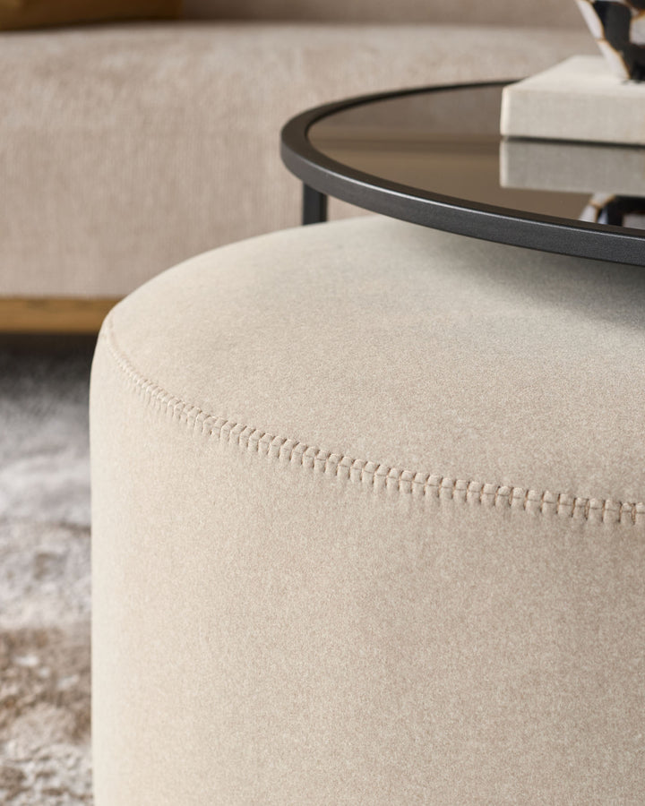 Cream upholstered cylindrical pouf with visible stitching detail, paired with a black round coffee table in a living room setting.