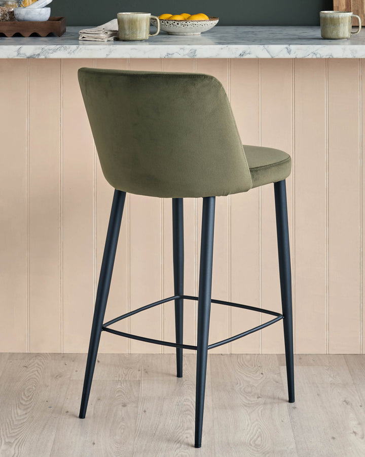 A green velvet bar stool with black metal legs sits in front of a counter, against a wall with vertical panels.