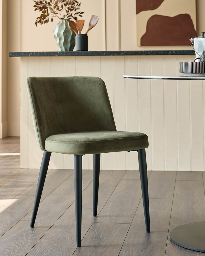 A stylish olive green dining chair with black legs sits on a wooden floor beside a round table, creating a modern aesthetic.