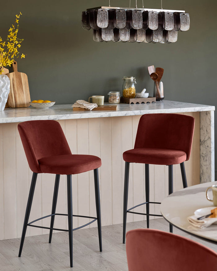 Stylish kitchen featuring two red velvet bar stools with black legs, alongside a round table and overhead chandelier.
