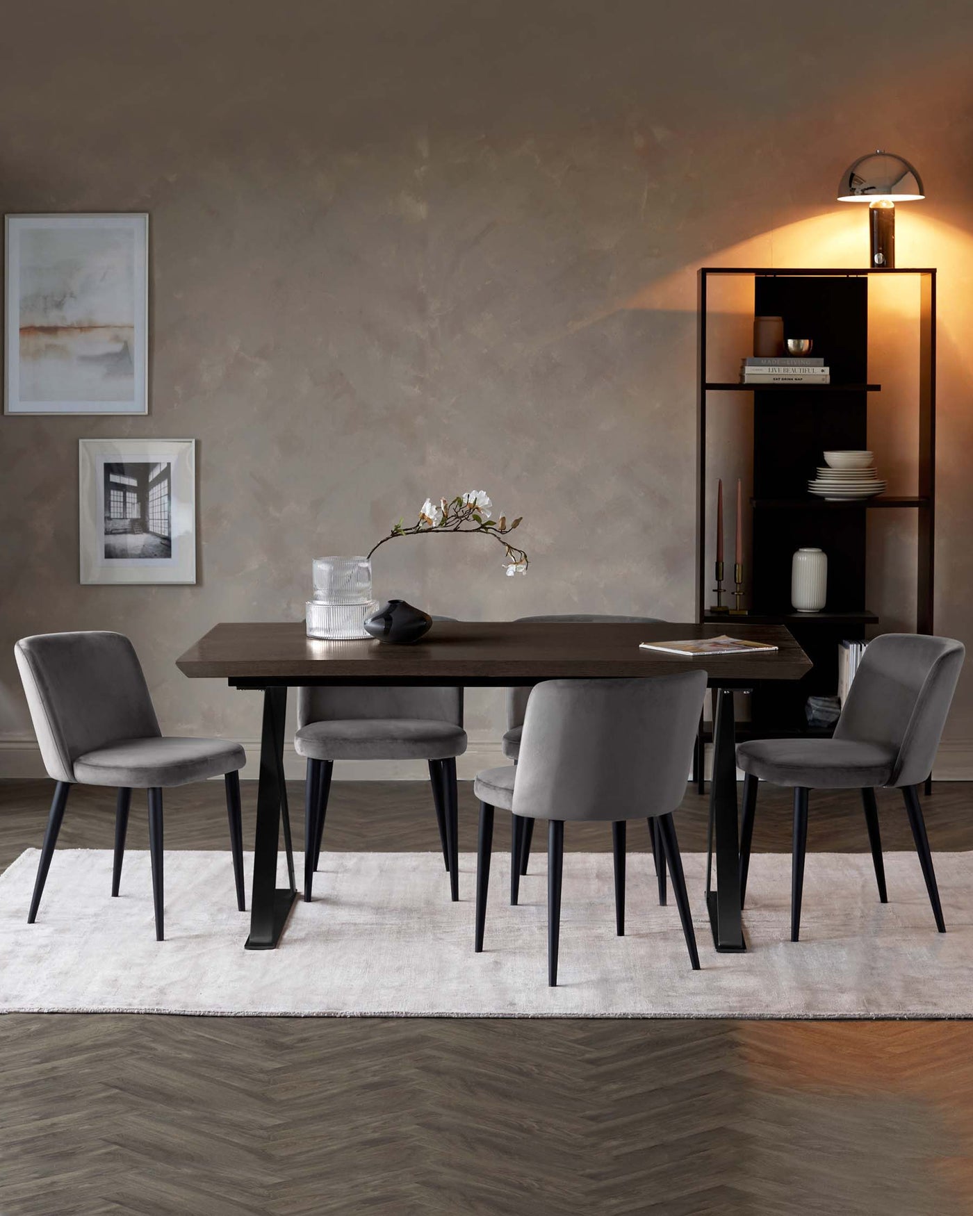 Contemporary dining room furniture featuring a dark wood table with angled black legs, paired with four plush, upholstered chairs in a light grey fabric and black slender legs. In the background, a black shelving unit with varying compartment sizes displays decorative items and books, illuminated by a stylish arching floor lamp with a chrome finish. The setting is complemented by a neutral rug under the table and chairs.