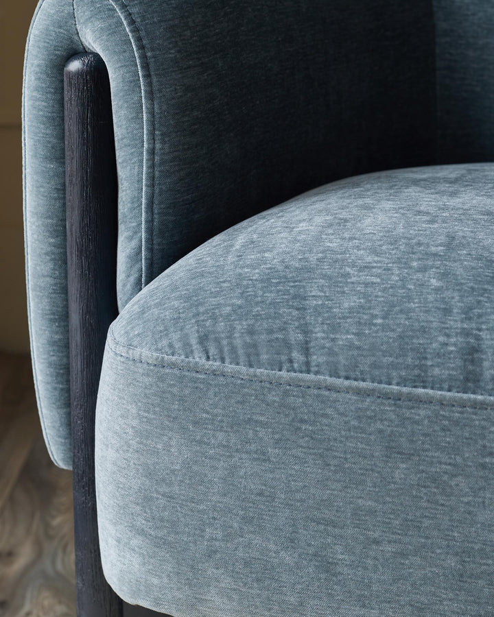 Close-up of a blue velvet armchair with a dark wooden armrest. The seat is rounded and has subtle stitching details.