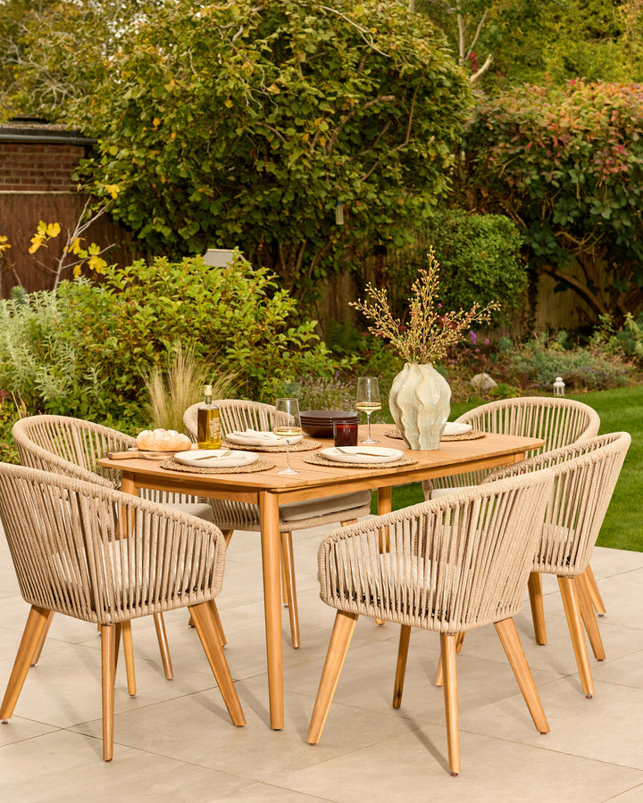 Tuscany teak wood 6-seater dining table with four woven rope chairs on a patio, styled with place settings and centerpiece.
