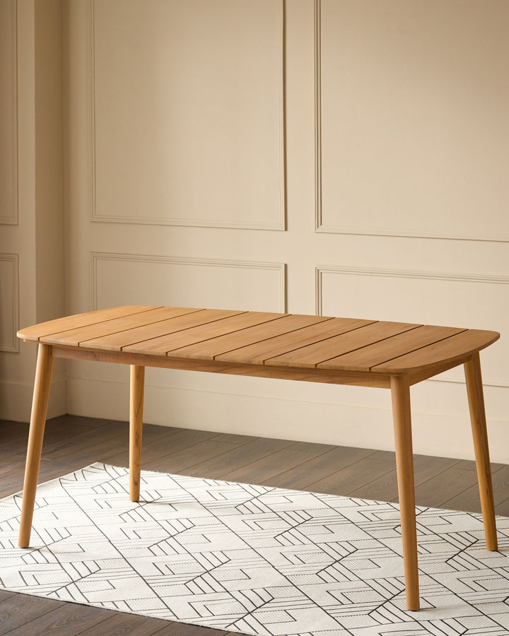 Tuscany teak wood dining table with slatted top, angled legs, shown in light room with geometric patterned rug.