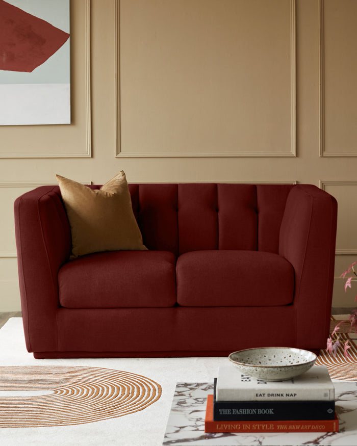 A plush, burgundy two-seater sofa with a gold cushion, atop a rug with a unique concentric circle pattern.