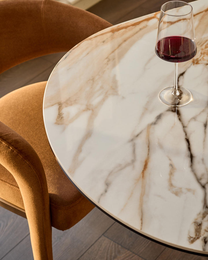 Vidal round dining table in natural ceramic with marble veining, paired with tan upholstered chair and red wine glass.