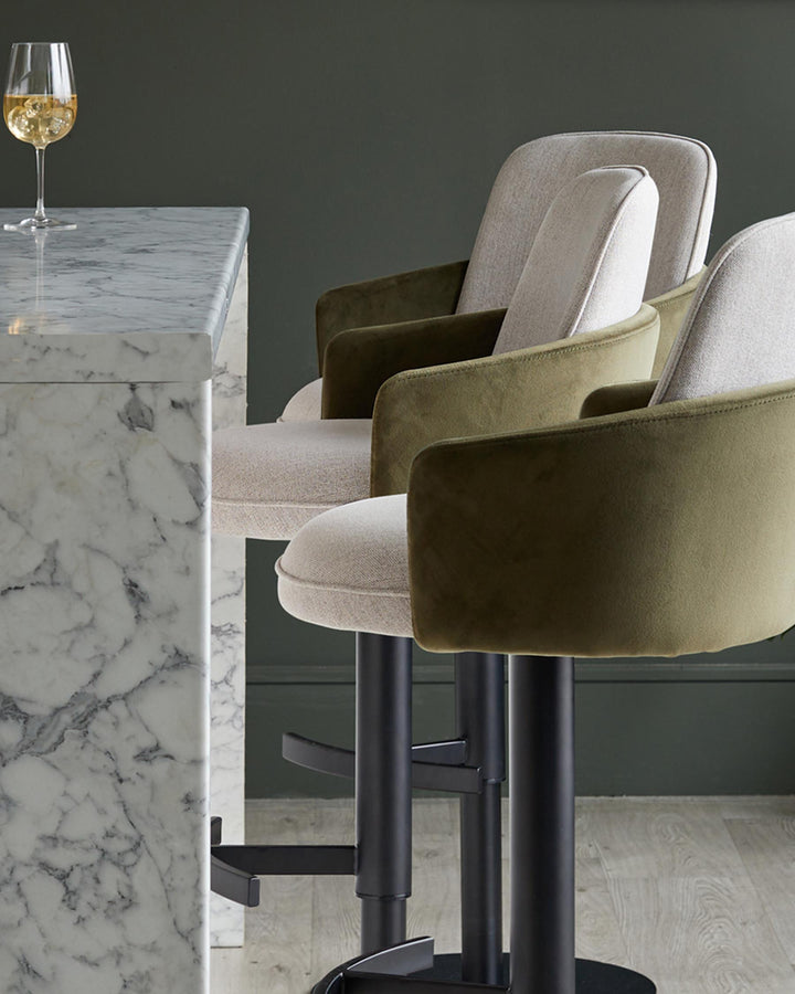 Contemporary bar stools with olive green velvet and cream fabric, plus a marble counter and wine glass.