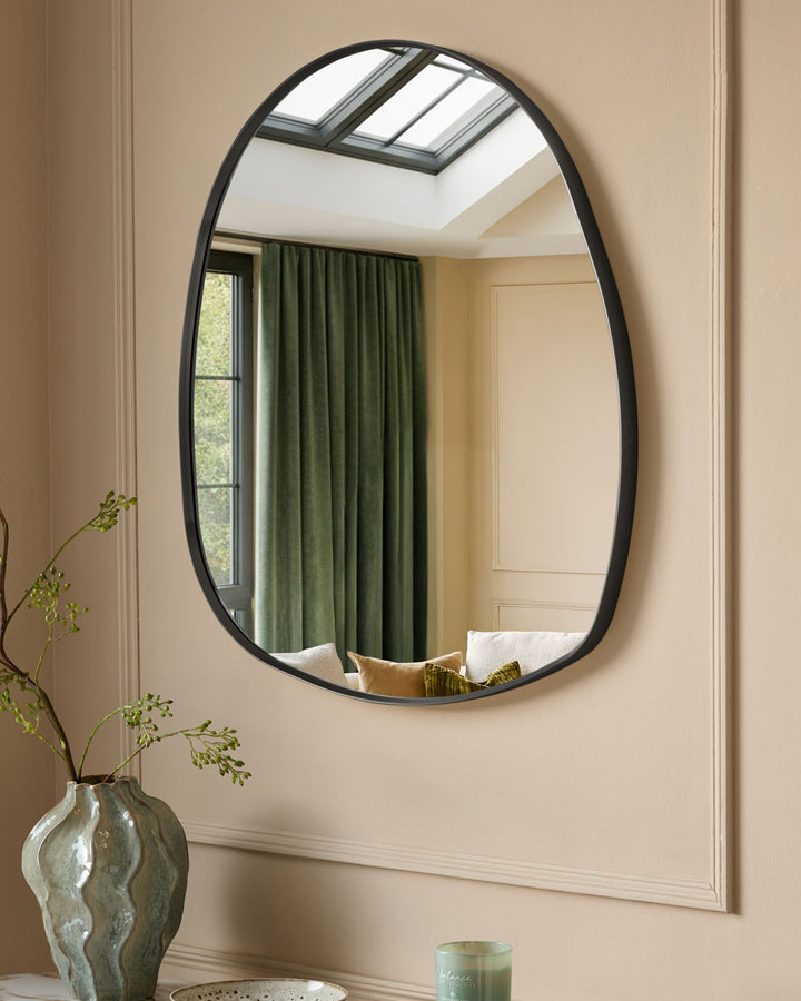 Black oval wall mirror with organic pebble-shaped frame, hung on beige wall above decorative vase with greenery.