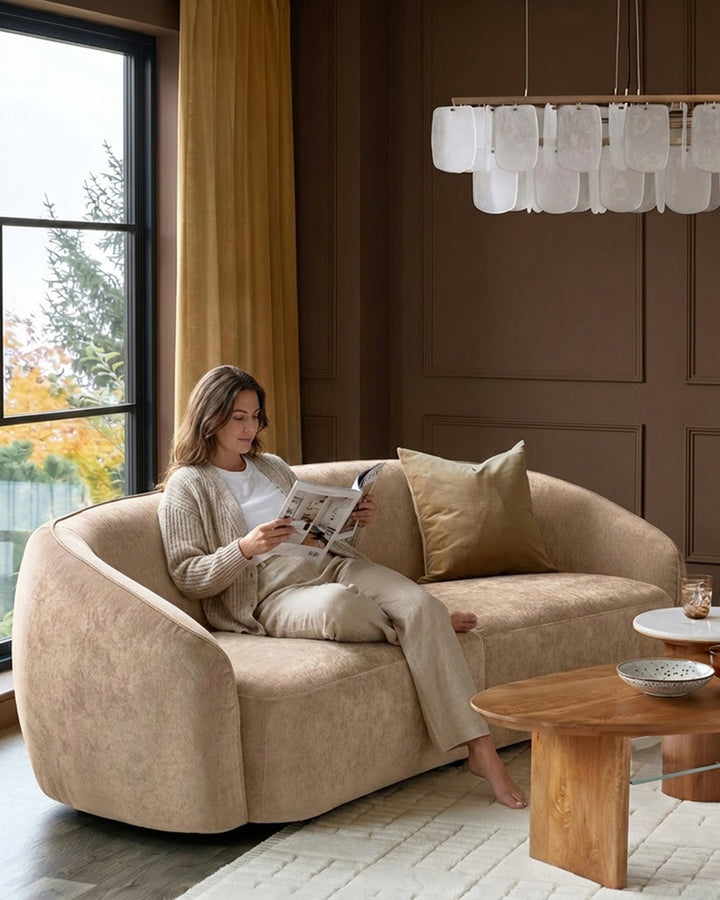 Zoey 3-seater sofa in champagne fabric with curved design, shown in a styled living room setting with a woman reading.