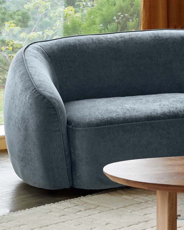 A modern, blue velvet sofa and a light wooden side table sit on a cream rug, beside a window with lush greenery.