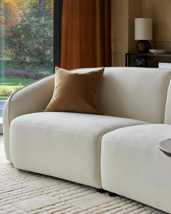 A modern cream sofa is pictured with a brown velvet cushion, alongside a lamp, table, and greenery.