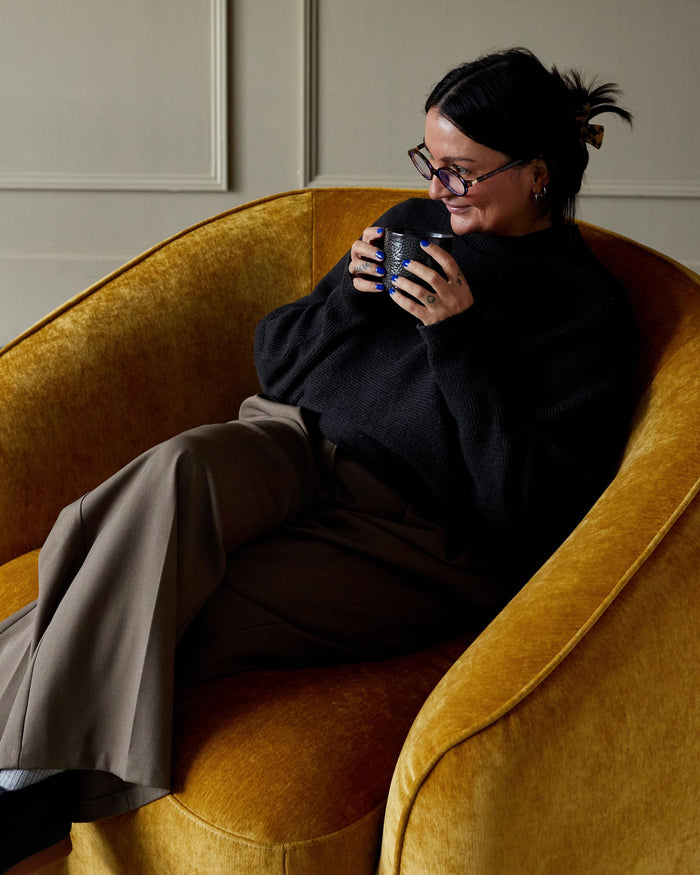 A woman sits in a yellow velvet armchair, holding a dark patterned mug. The chair has a curved back and a luxurious feel.