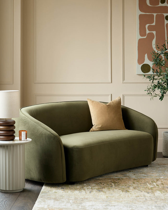 A stylish olive green loveseat with a beige cushion sits next to a ribbed side table and a decorative lamp.
