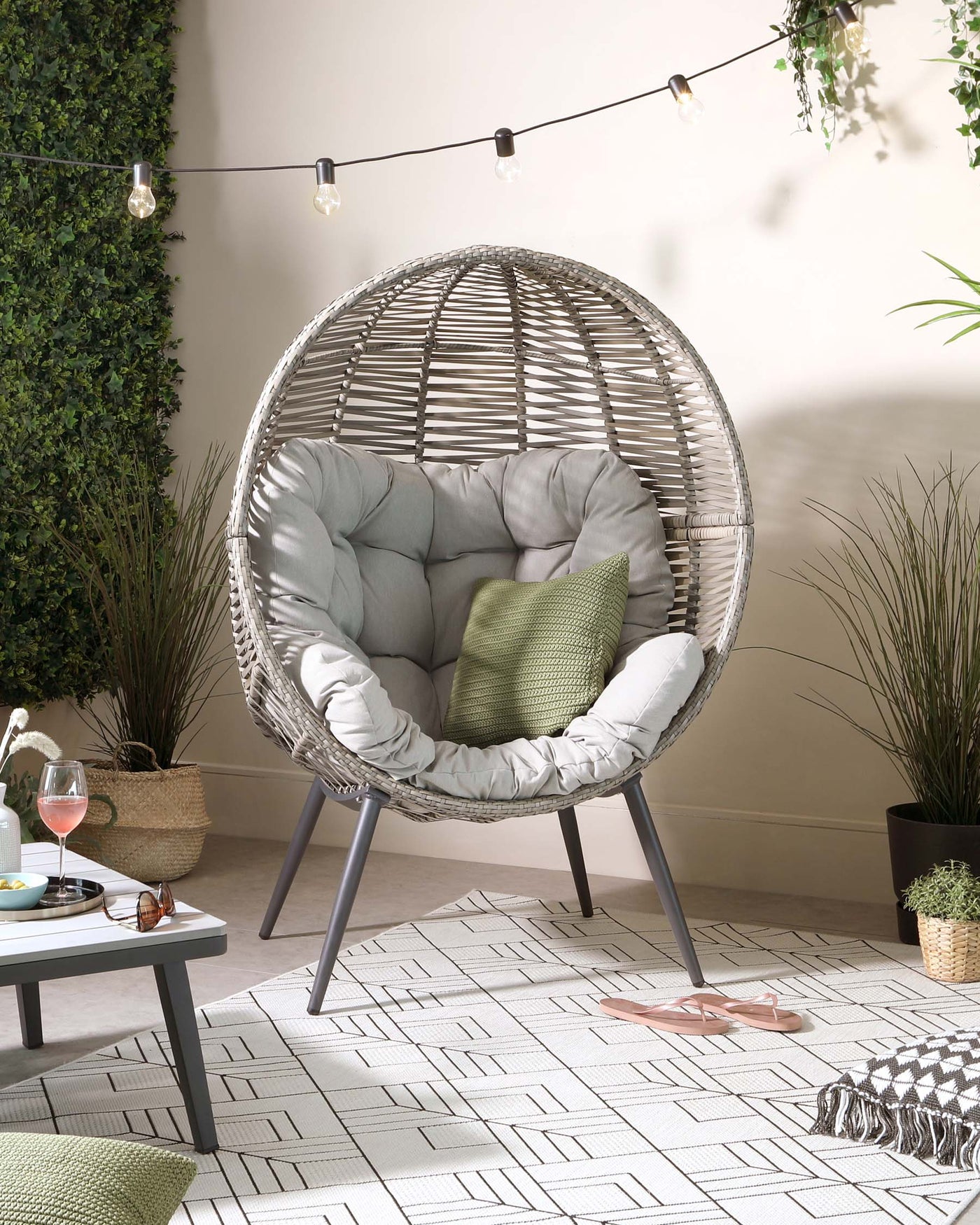 Egg-shaped wicker hanging chair with plush grey cushions, on a metal stand, complemented by a grey side table, placed on a patterned outdoor rug. The setting includes decorative outdoor string lights, green plants, and accent pillows, creating a cosy patio ambiance.