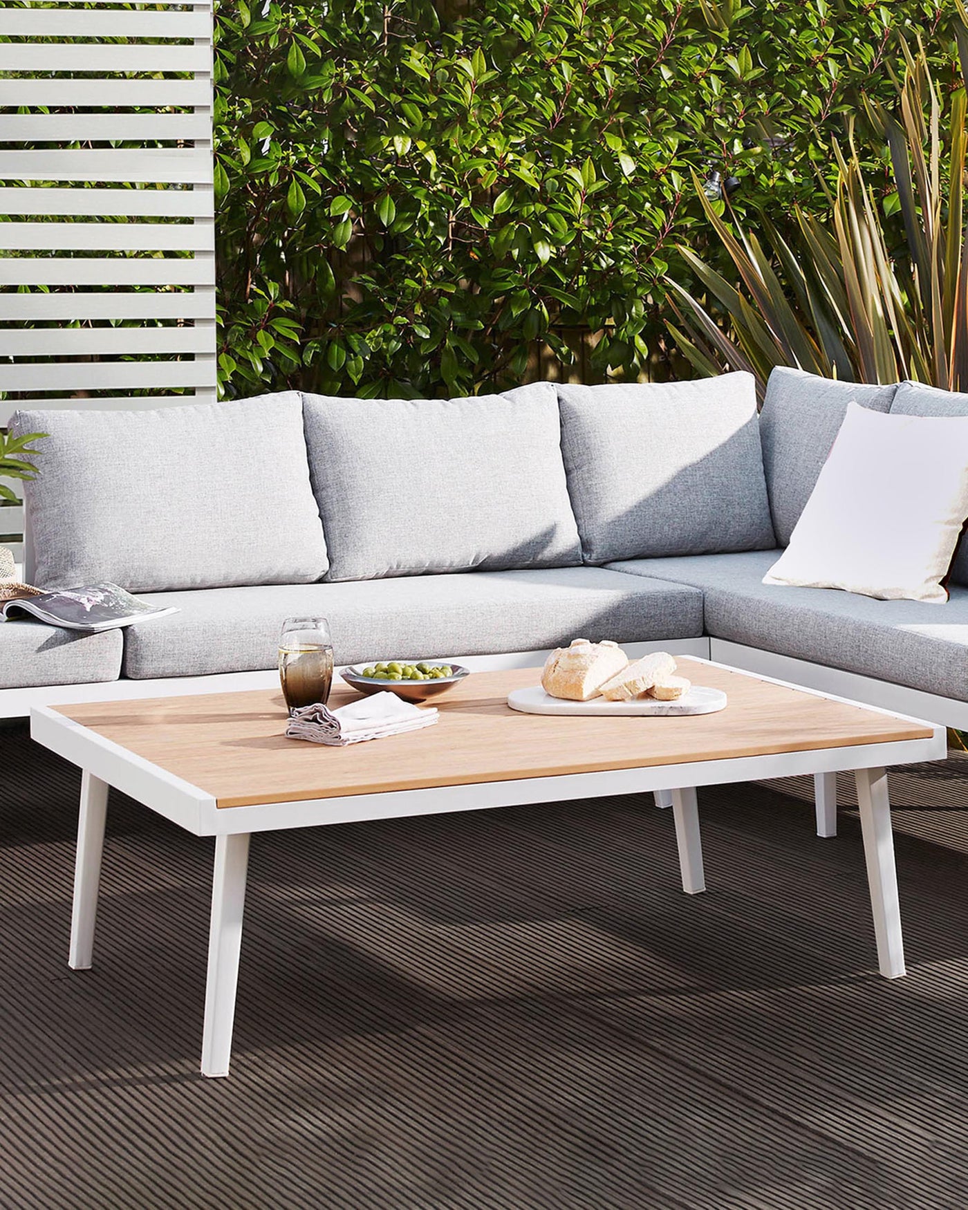 Modern outdoor furniture set consisting of a sectional corner sofa with light grey cushions and white throw pillows, accompanied by a rectangular coffee table with a white base and a wooden top. The set is showcased on an outdoor patio with a backdrop of green foliage.