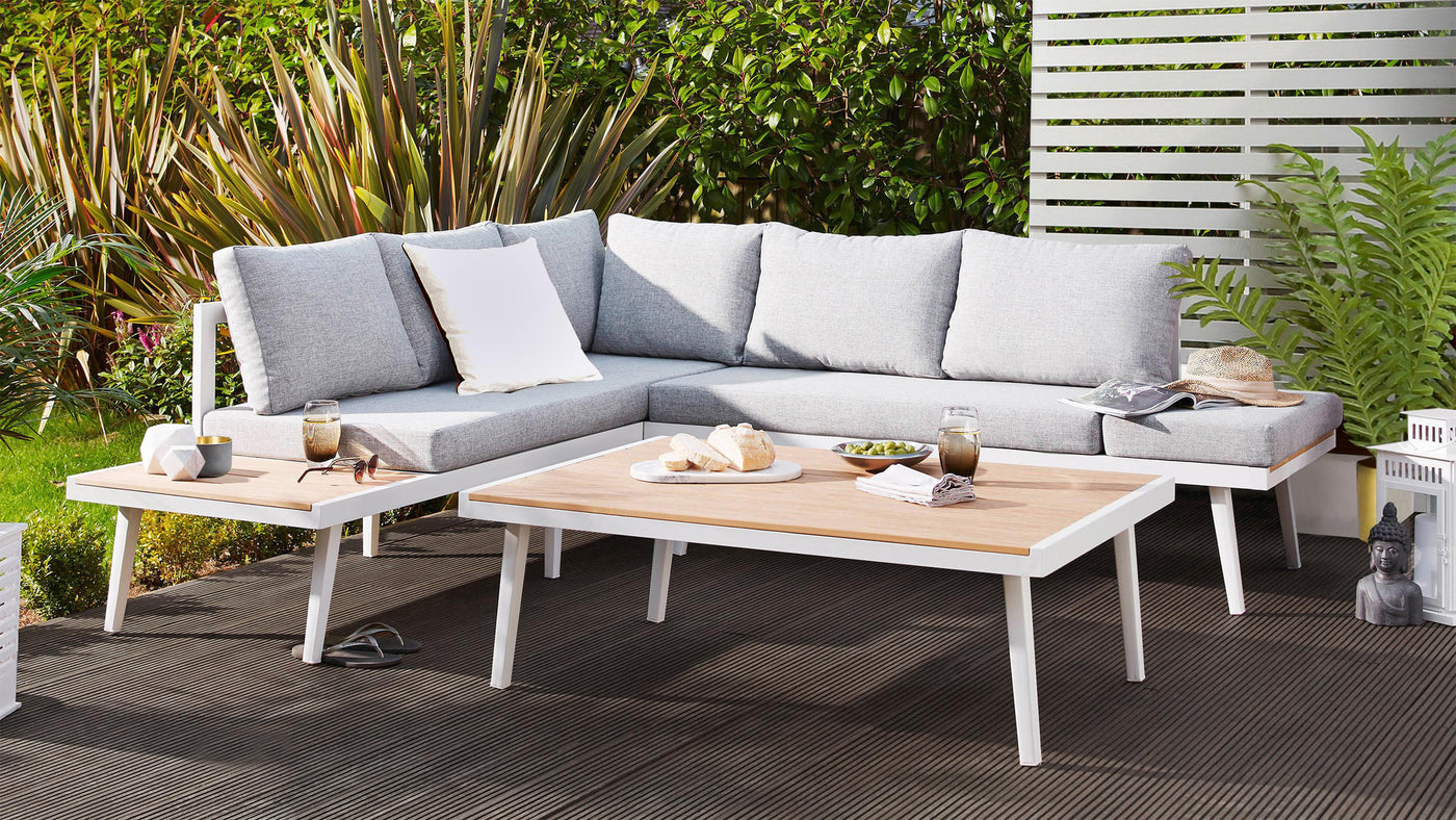 Modern outdoor furniture set on a patio, featuring a sleek, sectional corner sofa with light grey cushioning and white frames, paired with a white and natural wood rectangular coffee table.