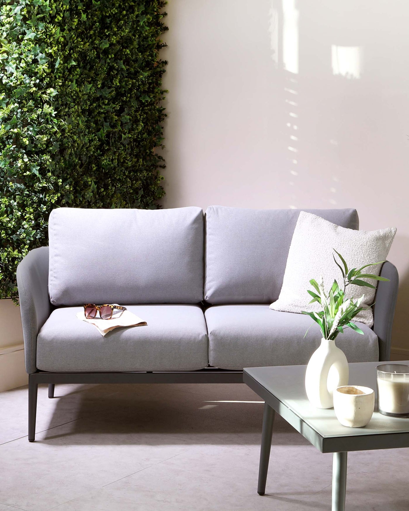 A modern minimalist style living room scene with a sleek grey upholstered two-seater sofa with clean lines, complemented by a light grey square coffee table made of a matte finish material, and accessorized with a white ceramic vase holding green foliage, alongside two candles on the tabletop. The backdrop features a vertical garden of lush greenery juxtaposed against a neutral wall, providing a refreshing contrast and a sense of tranquillity to the setting.
