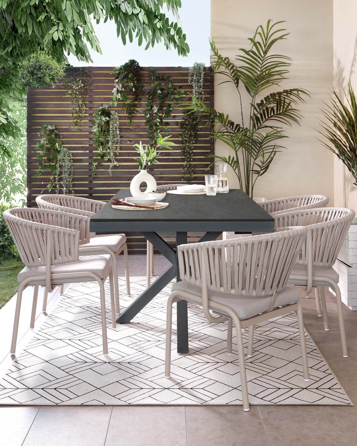 Outdoor dining set featuring a modern black rectangular table and six contemporary armchairs with woven backrests in a light grey finish, set on a geometric patterned area rug. The ensemble is displayed in a cosy patio setting with greenery.