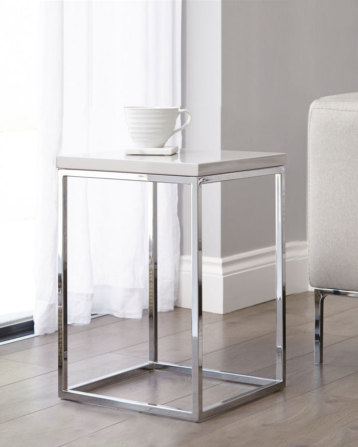 Modern minimalist side table with a white marble top and shiny chrome frame, featuring a square design and a sleek silhouette. A white ceramic cup and saucer rest on the table's surface, illustrating its use in a home setting.