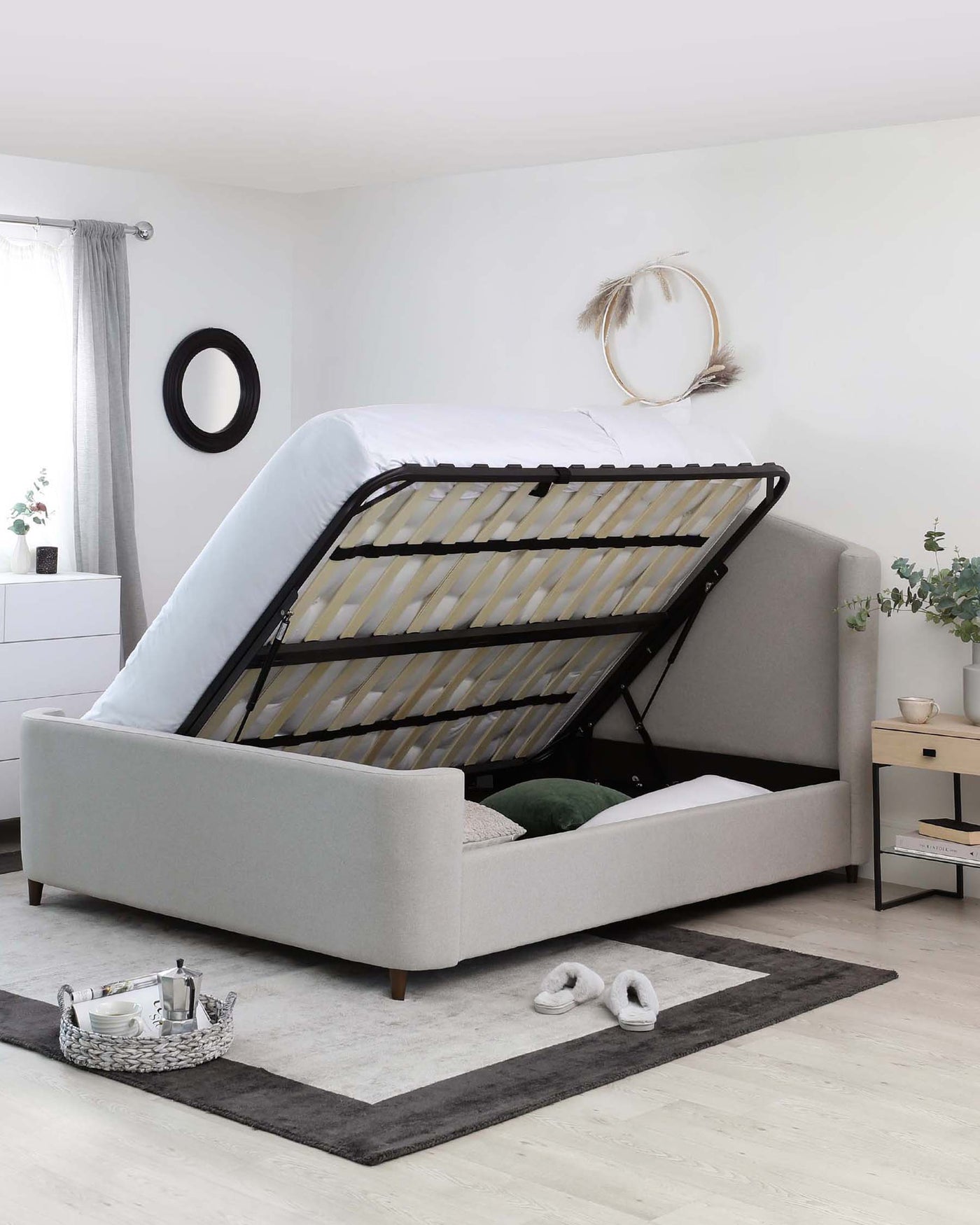 A modern light grey upholstered storage bed with a lifted mattress platform revealing an ample storage space underneath. The bed features a clean, streamlined design with a padded, fabric-covered frame and headboard, supported by four wooden legs. It's staged on a contemporary two-tone grey area rug over a light wooden floor, with minimalistic bedside tables on either side showcasing a simple, sleek white finish and handle less drawers.