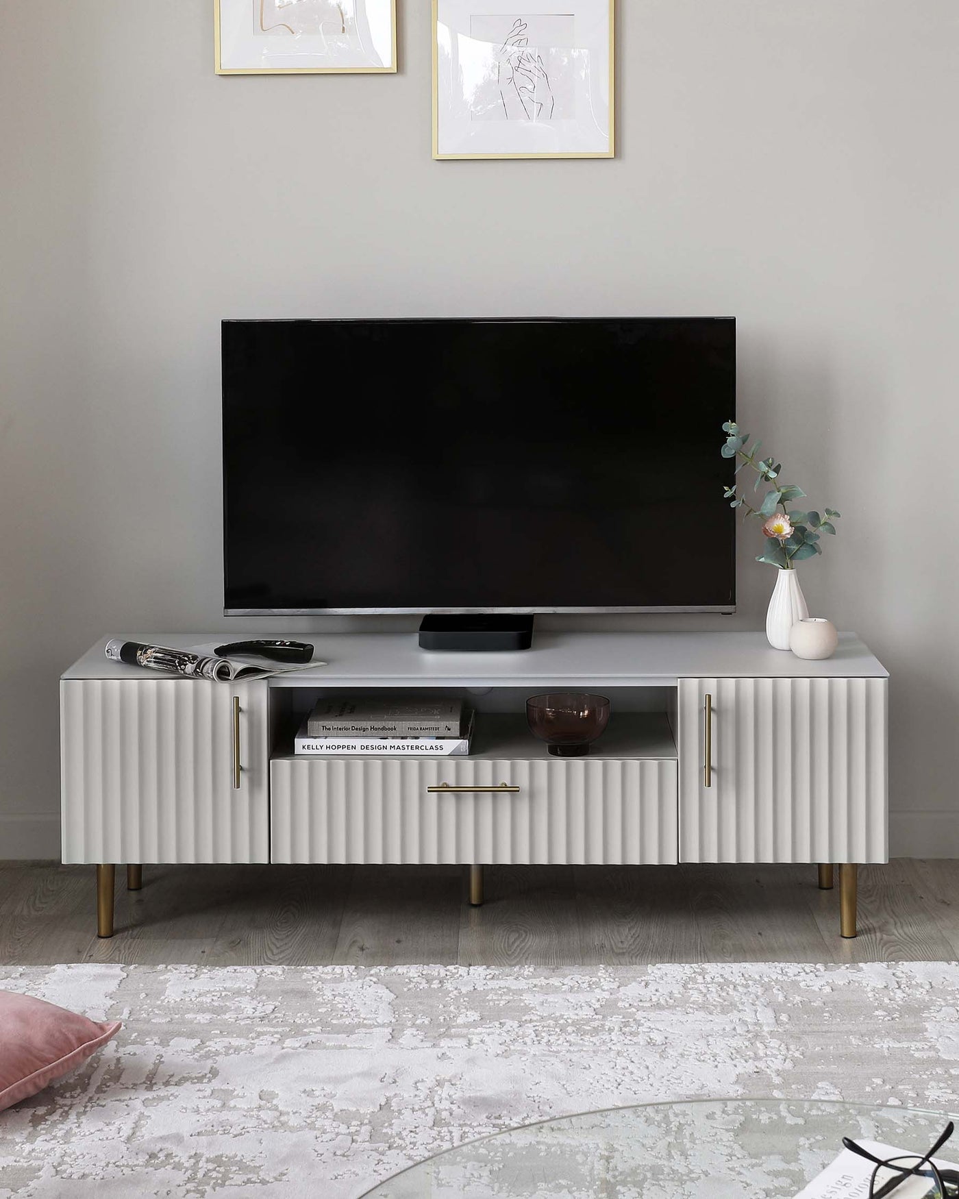 A modern white TV stand with textured front panels and gold-finished handles, featuring an open shelf and two compartments. The stand is supported by four angled wooden legs with gold-coloured tips matching the handles.