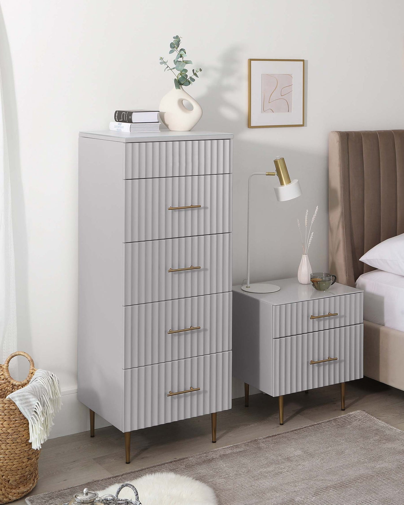 Modern bedroom furniture set featuring a tall, light grey, 5-drawer dresser with embossed front panels and brass handles, and a matching 3-drawer nightstand. Both pieces have splayed, tapered wooden legs.